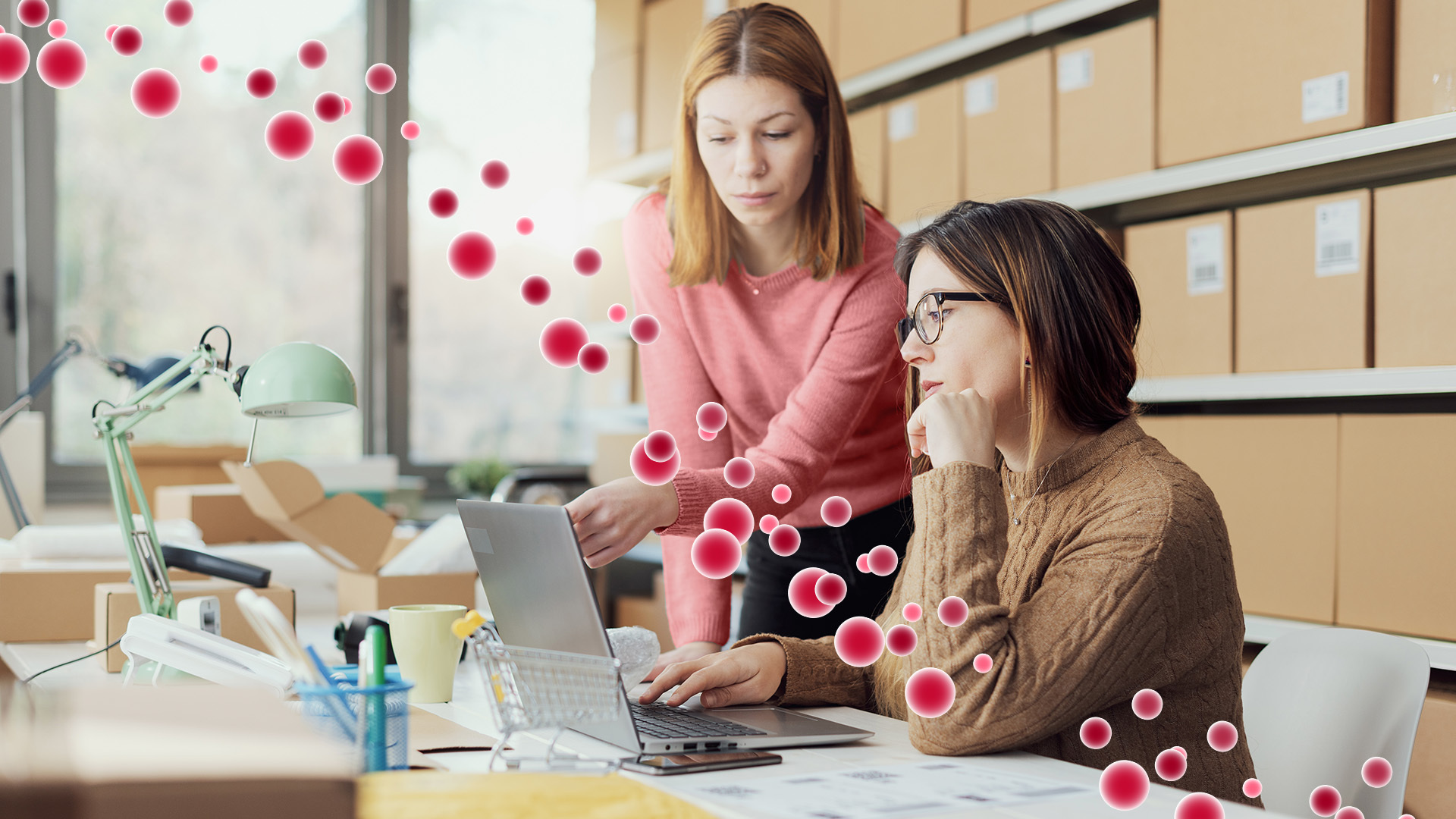 L'immagine rappresenta due donne lavorano al computer in ufficio, circondate da particelle rosse sospese nell’aria.